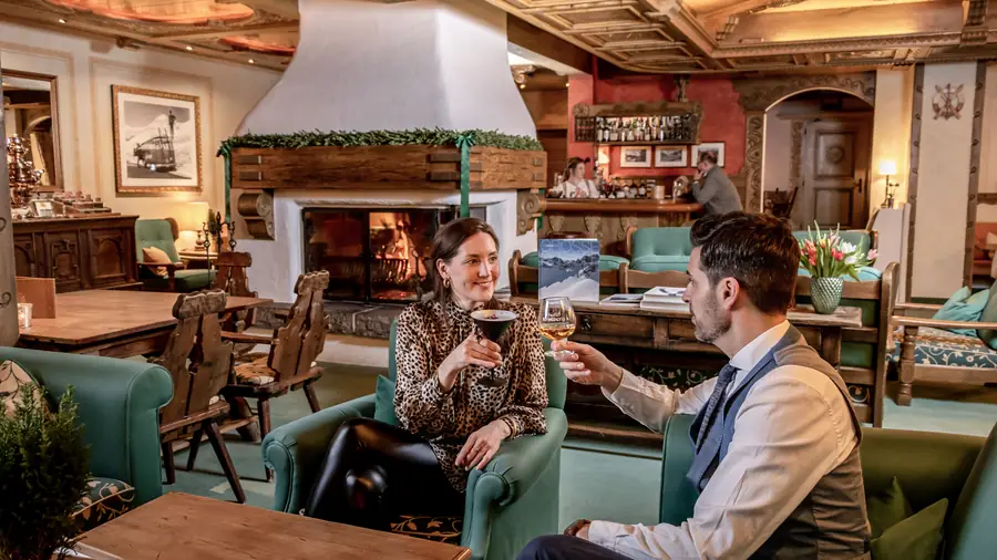 Kaminlounge mit offenem Feuer, zwei Gäste stoßen in grünen Sesseln an